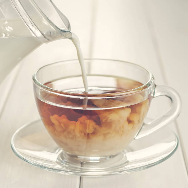 Pouring milk in tea in glass cup on white wooden table.