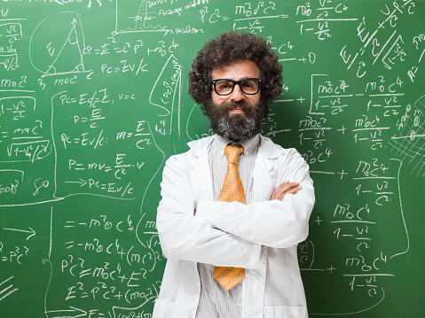 Portrait of mature adult teacher in front of green chalkboard. He is looking at camera and wearing a white lab coat and a yellow necktie.. Math formulas are written on blackboard. Shot in door with a medium format camera.