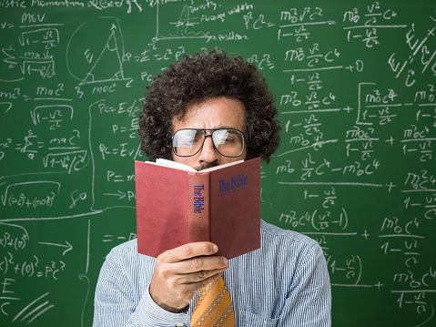 Mature adult scientist reading holy bible in front of green chalkboard. He has long brown curly hair and beard and is wearing a blue button down shirt with yellow necktie. There are mathematical formulas written on blackboard. Shot indoor with a medium format camera.