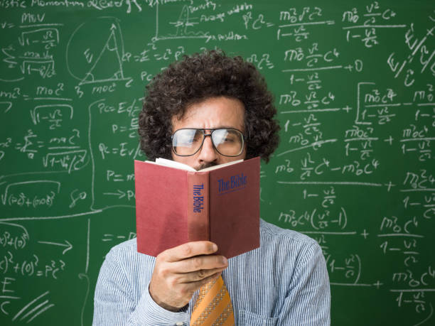 Portrait Of Mature Adult Scientist Reading Holy Bible Mature adult scientist reading holy bible in front of green chalkboard. He has long brown curly hair and beard and is wearing a blue button down shirt with yellow necktie. There are mathematical formulas written on blackboard. Shot indoor with a medium format camera. horn rimmed glasses stock pictures, royalty-free photos & images