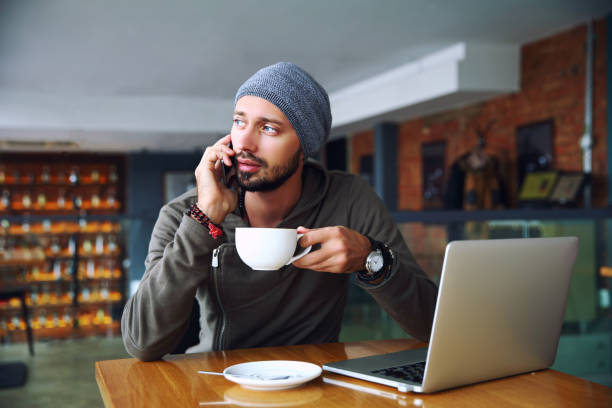 istock Young handsome hipster man with beard sitting in cafe talking mobile phone, holding cup of coffee. Laptop on wooden table 904348934