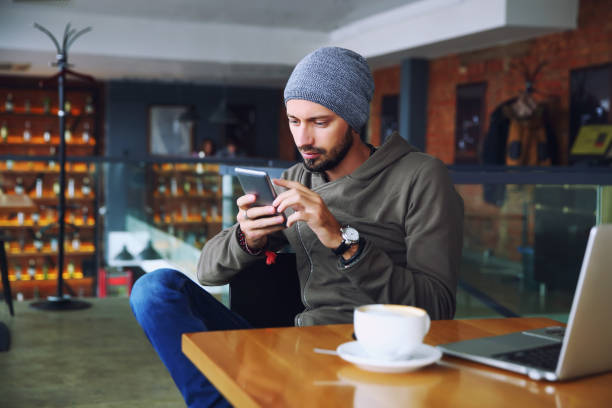 istock Young handsome hipster man with beard sitting in cafe talking mobile phone, holding cup of coffee and smiling. Laptop on wooden table 904351168