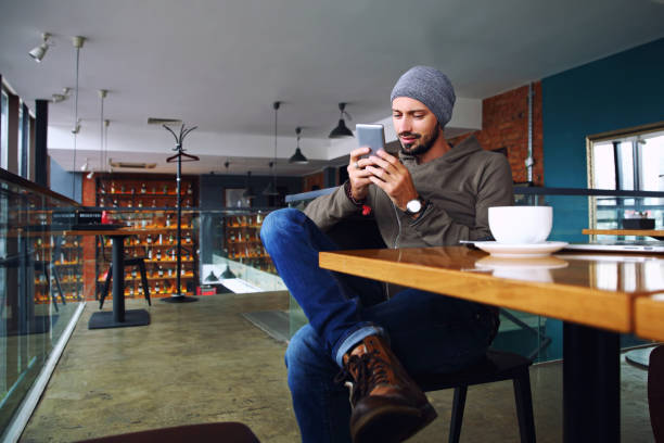 Young handsome hipster guy with beard texting with his mobile phone at the bar and having a cappuccino