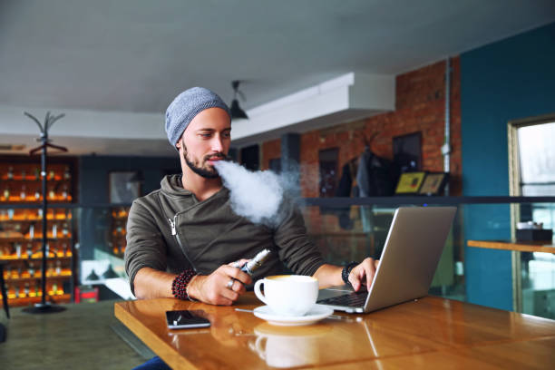 髭のリリース、vaping コーヒー カップでカフェに座っている若いハンサムなヒップスター男蒸気の雲。ノート パソコンで働いていて、少し休憩。コピー スペース - 電子タバコ ストックフォトと画像
