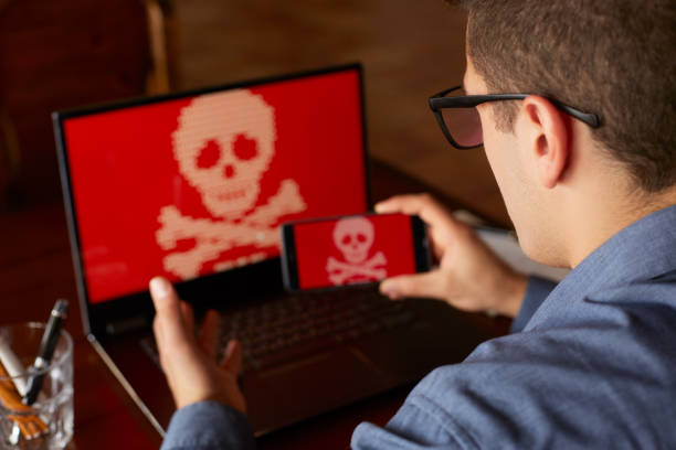 Man sits near laptop with phone blocked and encrypted by ransomware spyware asking for money. Laptop and smartphone infected by virus. Scary red skull crossbones on screen. Cyber security theme. hombre se encuentra cerca del ordenador portátil con el teléfono bloqueado y cifrado por software espía ransomware pidiendo dinero. portátil y smartphone infectados por virus. bandera pirata siniestro cráneo rojo en la pantalla. concepto de la seguridad de cyber