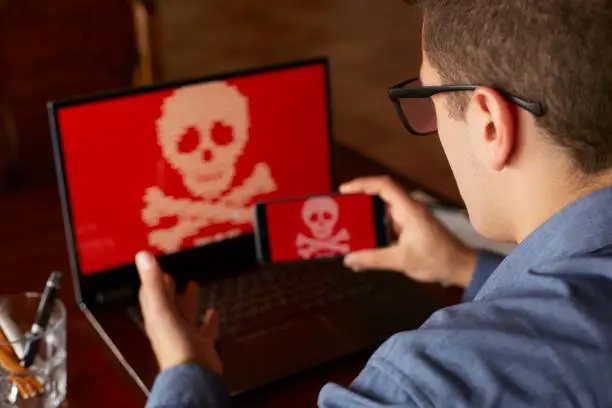 Photo of Man sits near laptop with phone blocked and encrypted by ransomware spyware asking for money. Laptop and smartphone infected by virus. Scary red skull crossbones on screen. Cyber security concept