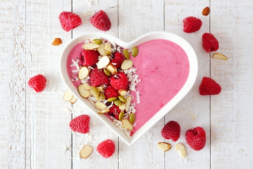 Healthy raspberry smoothie in a heart shaped bowl with superfoods. Above scene on a white wood background.