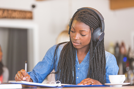 Young African American woman with long braided hair studies at a coffeehouse table while listening to music on her headphones. She is writing in her binder.
