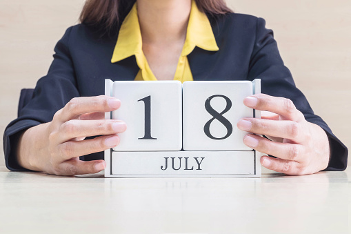 Closeup white wooden calendar with black 18 july word in blurred working woman hand on wood desk in office room , selective focus at the calendar