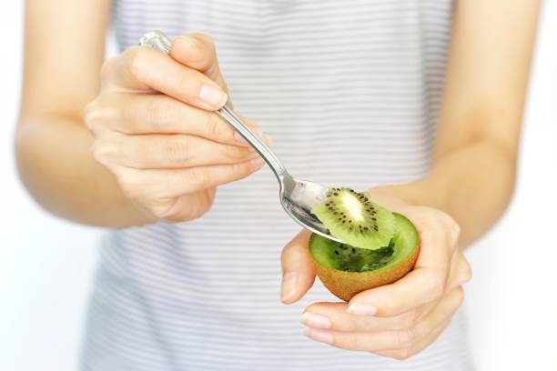 una donna tiene la frutta kiwi e affetta con il cucchiaio per mangiare su sfondo bianco isolato. concetto di nutrizione, salute, bellezza e dieta. - mature women isolated mature adult women foto e immagini stock