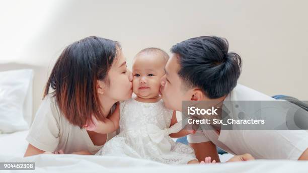Happy Family Young Asian Father And Mother Kissing Their Baby Girl On The Bedroom Stock Photo - Download Image Now