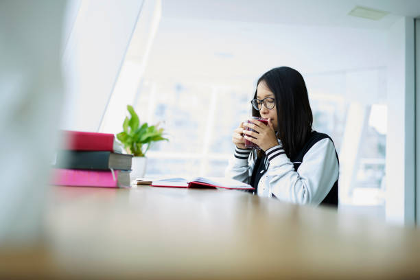 istock Beautiful asian young student casualy dressed in glasses for vision reading book and drinking tasty tea in university.Japanese teenager female dressed in bomber jacket preparing for exam 904460740