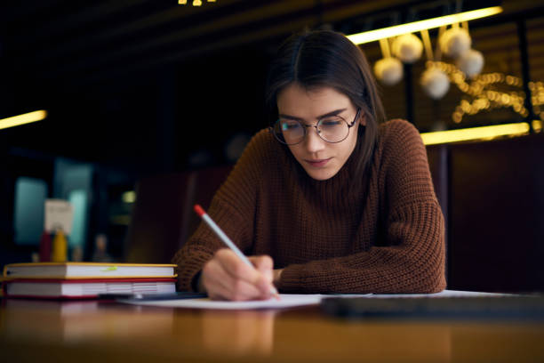 istock Pensive young female student in glasses working on coursework project preparing graphic planning and sketches sitting in coworking office with textbooks and equipment concentrated on fulfilling task 904463858