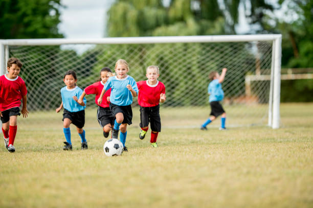 Chasing The Ball A group of elementary school kids are playing soccer together. They are wearing red and blue uniforms. They are chasing after the ball together. football league stock pictures, royalty-free photos & images