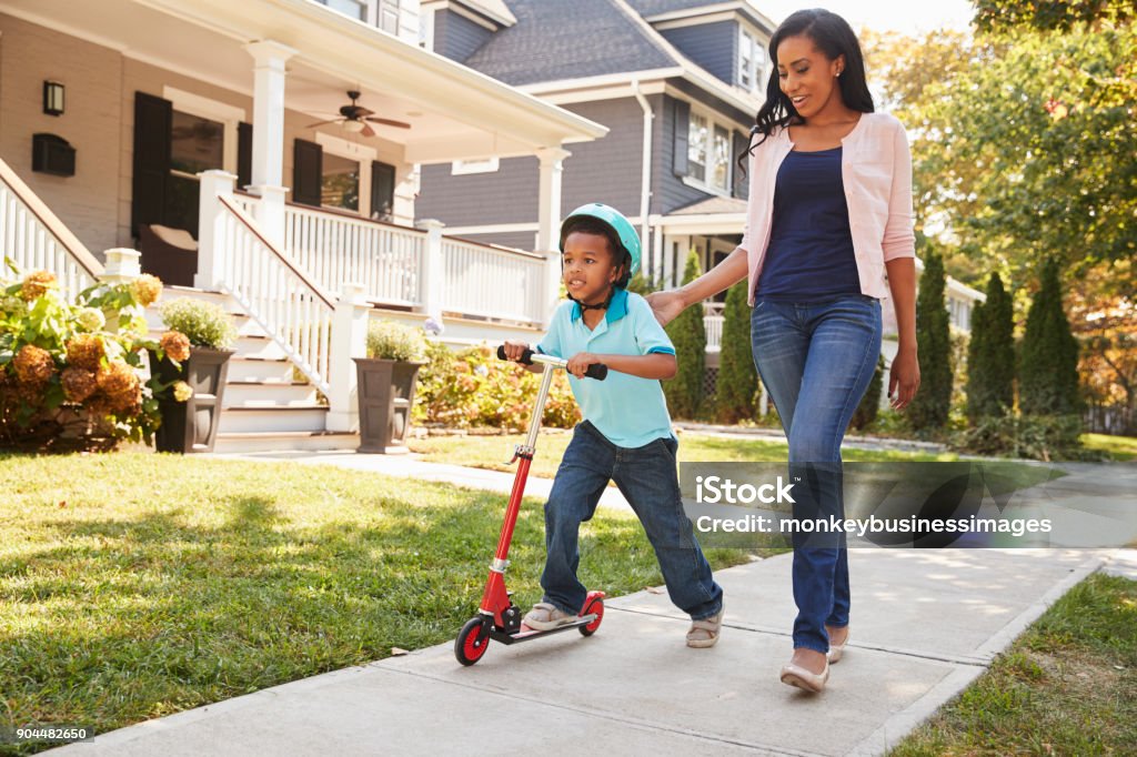 Mother Walks With Son As He Rides Scooter Along Sidewalk Family Stock Photo