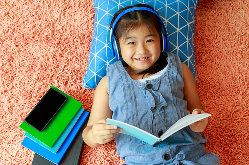 Happy Asian little girl laying down on floor with reading book and headphones listen music from mobile phone