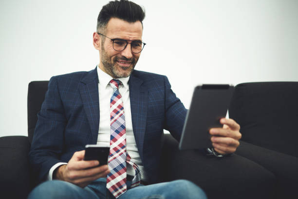 istock Handsome mature male executive manager in formal wear testing work of innovative software for digital devices updating application on smartphone and touch pad using wifi connection in coworking office 904510396