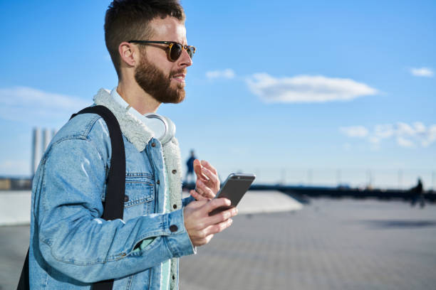 istock Young handsome bearded guy strolling on street using application on smartphone, confident male hipster dressed in jeans jacket and sunglasses holding cellular sending messages using 3G internet 904514174