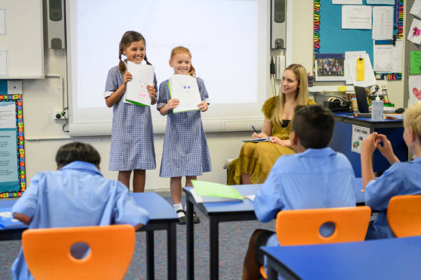 Two school girls presenting their work to the class with their teacher watching