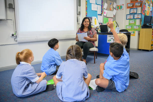 School children sitting on classroom floor listening to their teacher School boy with hand up answering the teacher's question at school kid sitting cross legged stock pictures, royalty-free photos & images