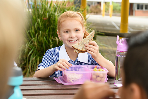 Cheerful school pupil sitting outdoors with packed lunch and eating healthy food