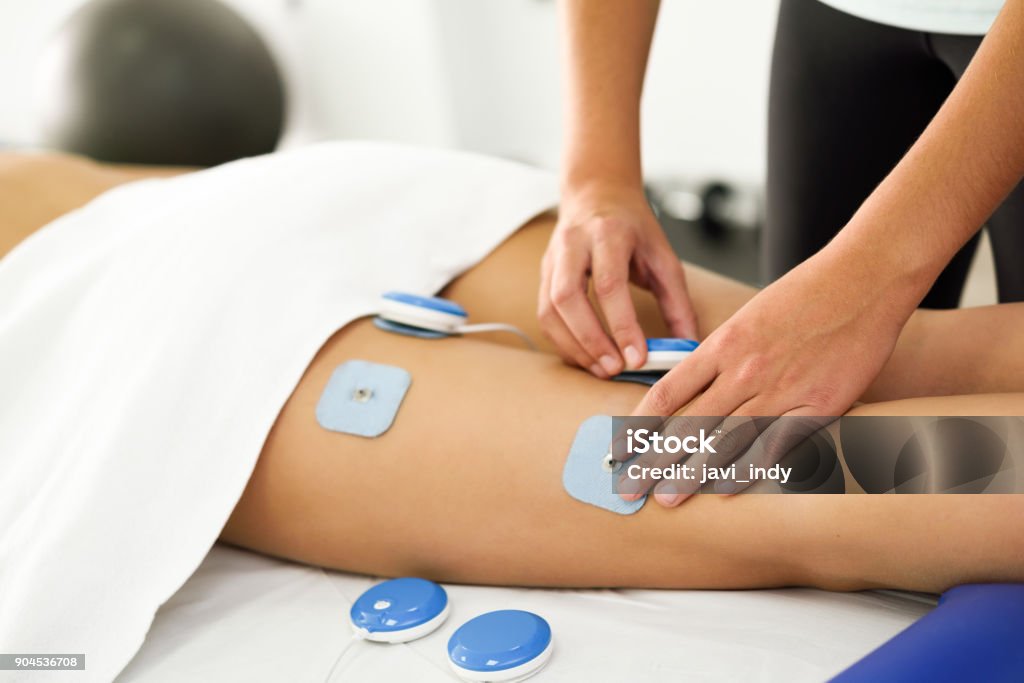 Physiotherapist applying electro stimulation in physical therapy to a young woman leg. Physiotherapist applying electro stimulation in physical therapy to a young woman. Medical check at the leg in a physiotherapy center. Adult Stock Photo