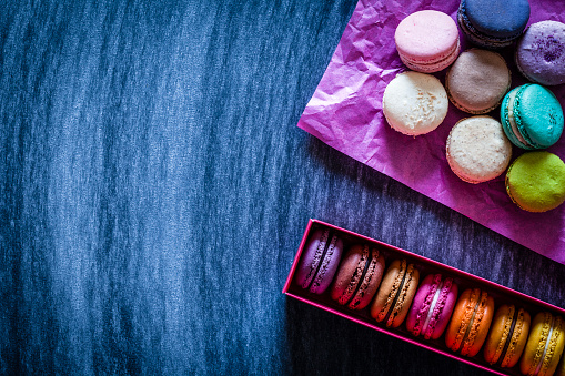Multi colored macaroons arranged in a box shot from above on blue table. Some macaroons are out of the box placed directly on the table on pink paper napkin. Low key DSRL studio photo taken with Canon EOS 5D Mk II and Canon EF 100mm f/2.8L Macro IS USM