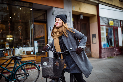 Smiling young woman riding bicycle. Female is cycling on street by building. She is in warm clothing at city.