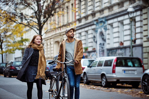 Friends walking with bicycle on street. Man and woman are in warm clothing. They are traveling together in city during winter.