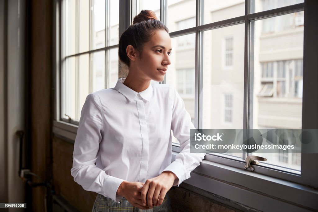 Young woman with hair bun looking out of window, waist up Women Stock Photo