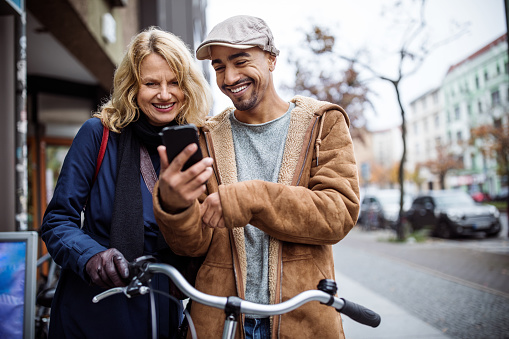 Happy man showing mobile phone to friend. Male and female are standing with bicycle. They are in warm clothing at city.