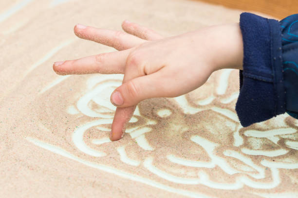 painting, creativity, handmade concept. close up of little arm of kid dressed in denim shirt, he is drawing lines and curles with his finger on the white surface covered by sand stock photo