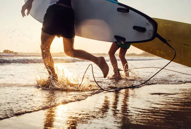 Photo of Son and father surfers run in ocean waves with long boards. Close up splashes and legs image