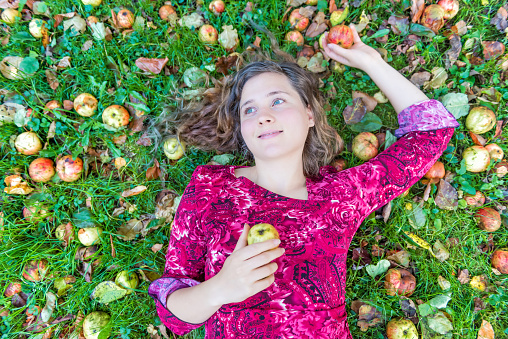 Smiling young woman girl face lying on ground with many apples fallen wild fresh on grass ground bruised on apple picking farm closeup
