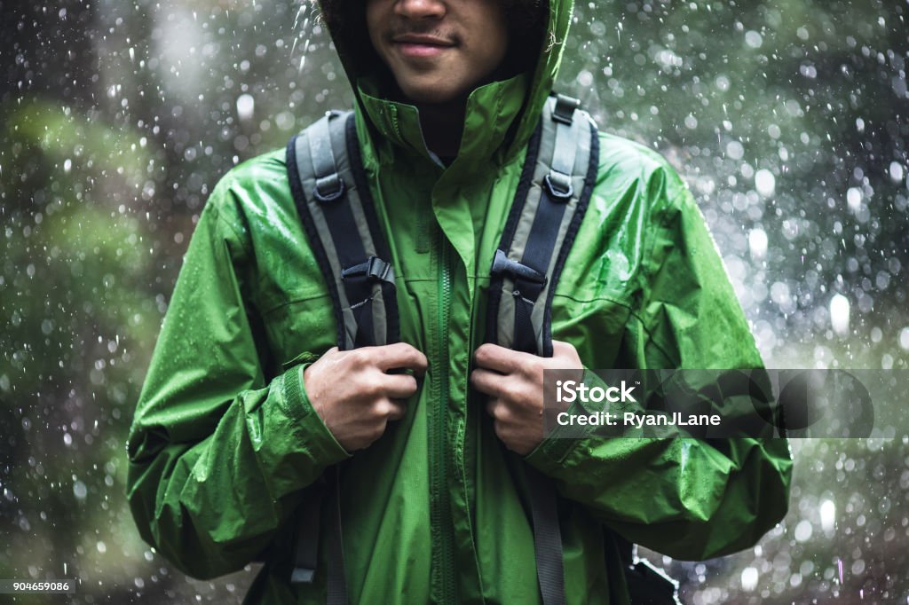 Young Man Hiking in Rain with Waterproof Jacket A cheerful young adult African American man goes for a hike in the rain in the Pacific Northwest, the raindrops repelling from his raincoat.  Shot in Washington state. Hiking Stock Photo
