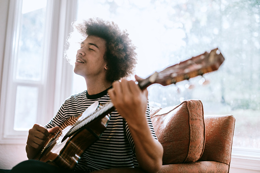 A young adult African American man plays an acoustic guitar in his home, the sun light streaming through the window as he enjoys playing and singing with his instrument.