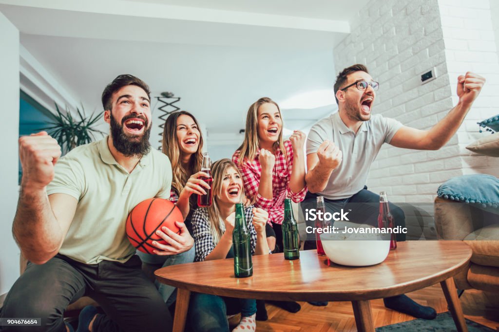 Group of friends watching a basketball game on tv Happy friends or basketball fans watching basketball game on tv and celebrating victory at home.Friendship, sports and entertainment concept. Apartment Stock Photo