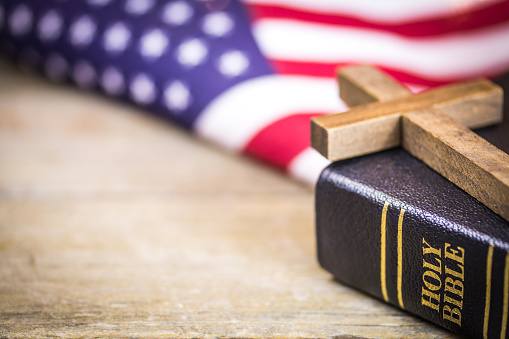 A wooden Christian cross laying on a holy Bible with an American flag background.