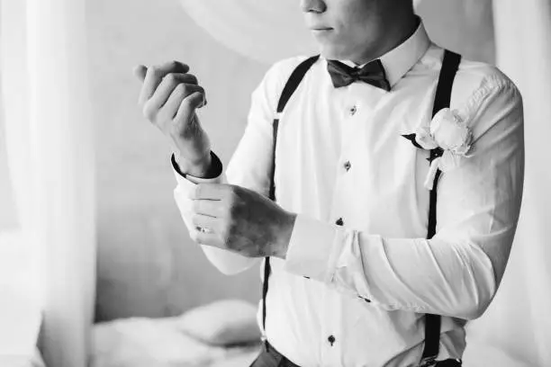 Photo of Grooms morning preparation handsome groom in bow tie suspenders getting dressed and preparing for the wedding at hotel the bridegroom wears cufflinks in a white shirt black and white portrait