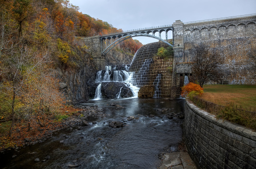 The New Croton Dam water falls and the road over the top with a flowing creek in foreground.Fall vegetation on either side.