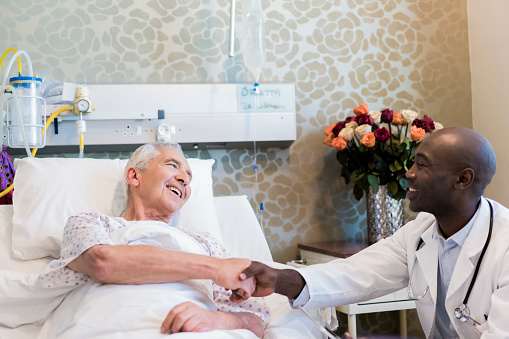 Smiling doctor shaking hands with senior patient. Practitioner sitting by man lying on bed. Multi-ethnic males are in hospital ward.