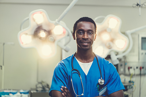 Portrait of smiling male doctor against illuminated lights. Young healthcare worker is wearing blue scrubs. He is standing with arms crossed in hospital.