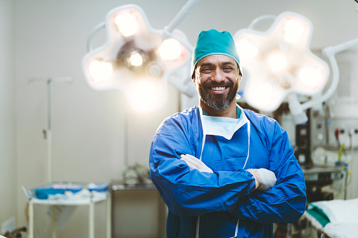 Portrait of smiling surgeon in hospital. Male healthcare worker is wearing scrubs. He is standing with arms crossed against lights.