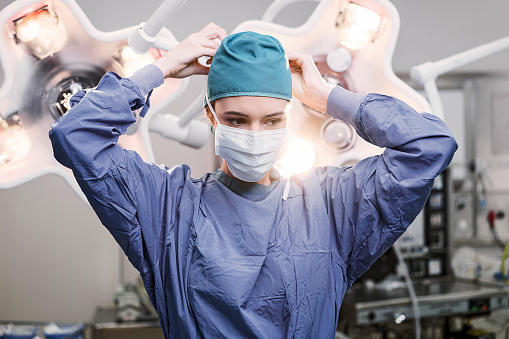 Confident female doctor tying surgical mask. Healthcare worker is wearing scrubs. She is standing in operating room at hospital.