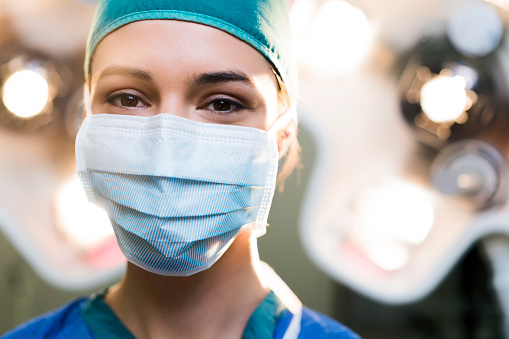 Portrait of female doctor wearing surgical mask. Confident healthcare worker is against illuminated lights. She is in operating room at hospital.