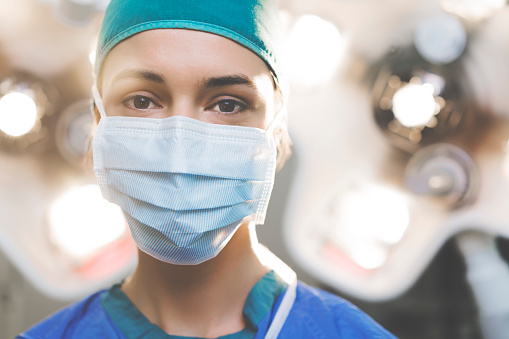 Portrait of confident female surgeon. Healthcare worker is wearing surgical mask. She is against illuminated lights in operating room at hospital.