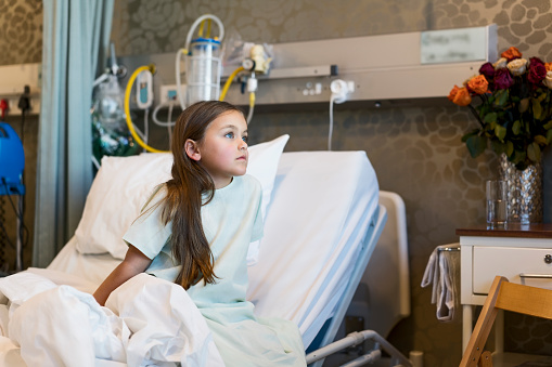 Thoughtful patient sitting on bed. Girl is in hospital gown at ward. She is looking away.