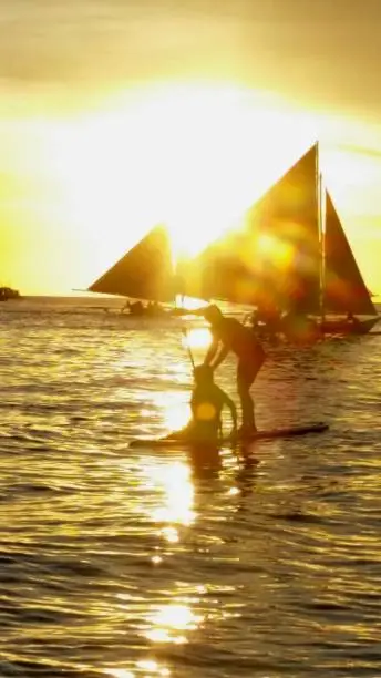 Two girls try to keep balance on a paddleboard on the beaches of boracay, Philippines while sailboats float by during the sunset.