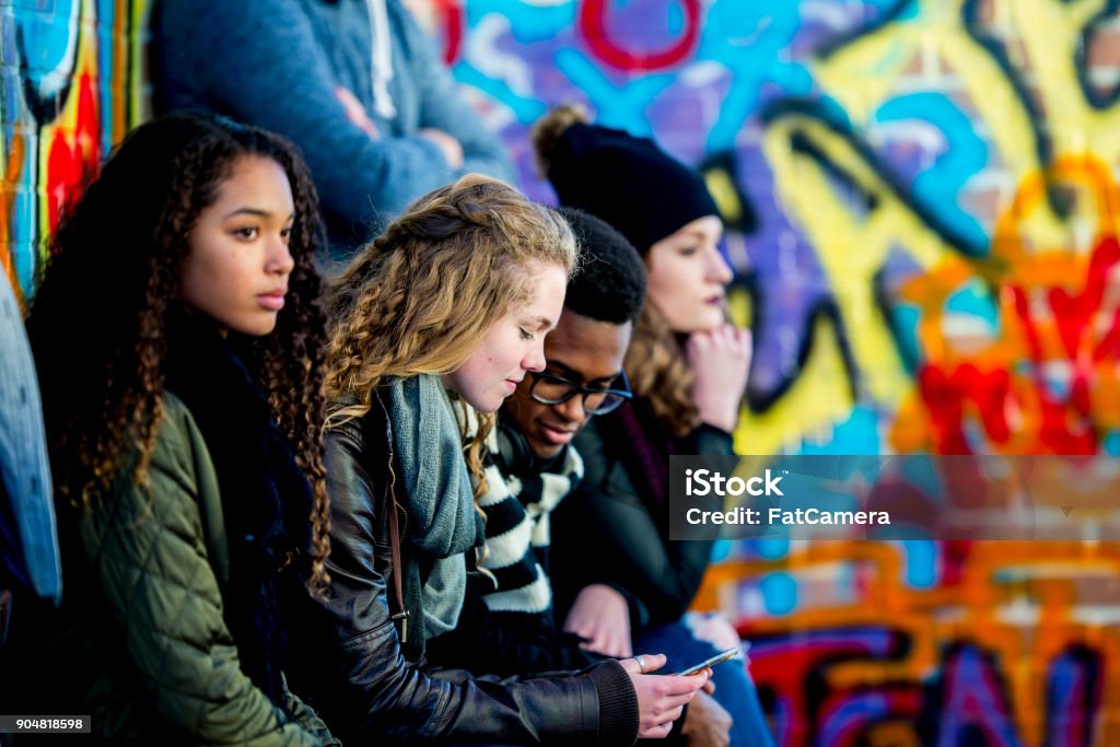 Teens With Technology A group of teenagers are sitting in front of a wall covered in graffiti. They are wearing stylish clothes. A boy and girl are looking at a smartphone screen together. Teenager Stock Photo