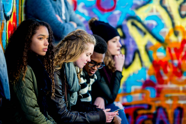 Teens With Technology A group of teenagers are sitting in front of a wall covered in graffiti. They are wearing stylish clothes. A boy and girl are looking at a smartphone screen together. teenagers only photos stock pictures, royalty-free photos & images